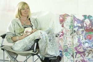 A woman sitting in a folding chair next to colorful oil paintings