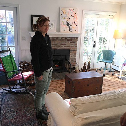 An artist stands in front of a fireplace and rocking chair in her studio