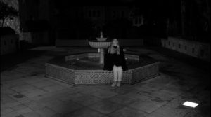 A black and white still of a girl standing in front of a fountain at night