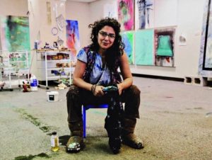 An artist sits on a stool in her studio with tools, with her paintings in the background