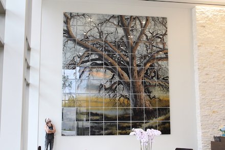An artist stands with a large-scale installation of a detailed painting of a dead tree