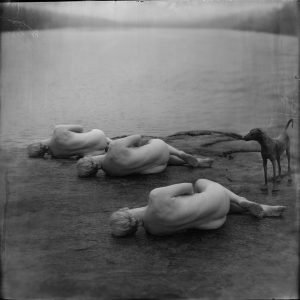 A black and white photograph of three naked women and a dog laying on the shore of a lake
