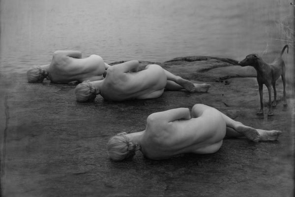 A black and white photograph of three naked women and a dog laying on the shore of a lake