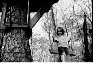 A black and white photograph of a young girl on a tree swing