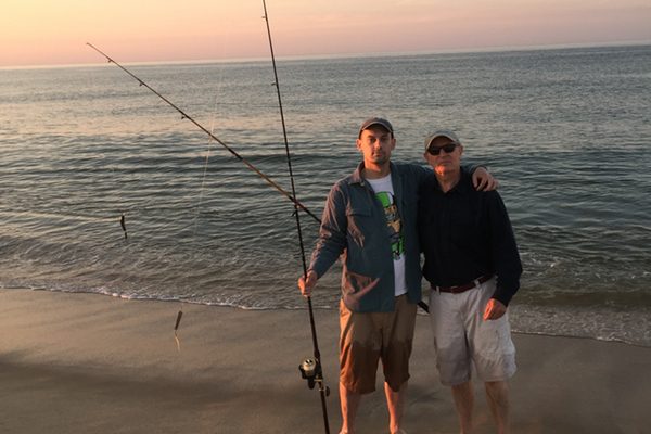 Two men stand on the ocean shore with fishing poles