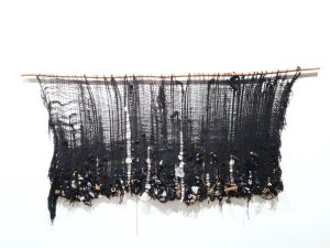 A black wall weaving with object woven into the bottom of the piece