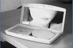 A black-and-white photograph of a woman looking into the mirror of a makeup compact