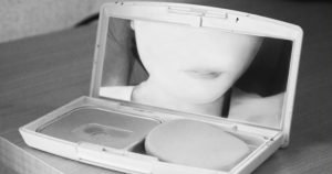 A black-and-white photograph of a woman looking into the mirror of a makeup compact