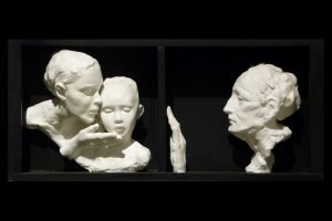 White bust statues of a woman whispering to a child, and an older woman watching