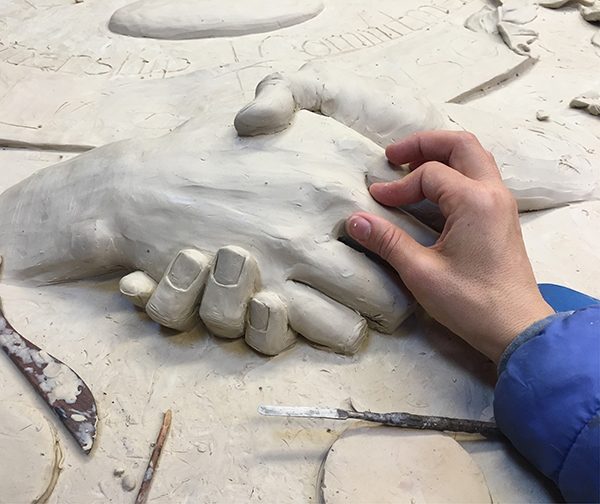 An artist works on shaping a clay sculpture of two hands shaking