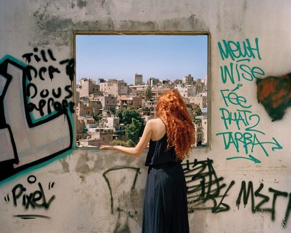 A photo of a woman looking out of a graffitied window overlooking a city