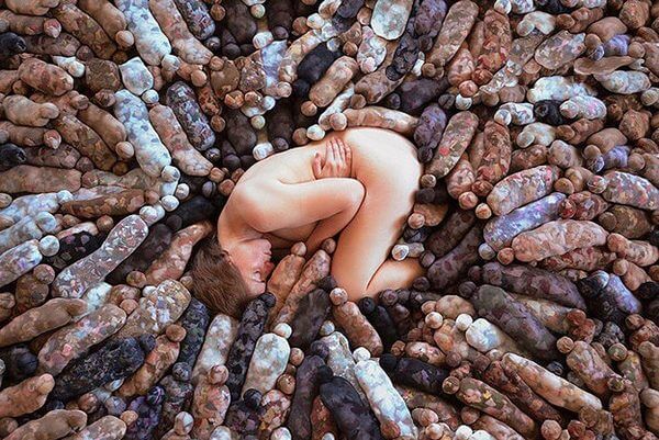 A photograph of a naked woman sleeping among a crowd of fabric dolls