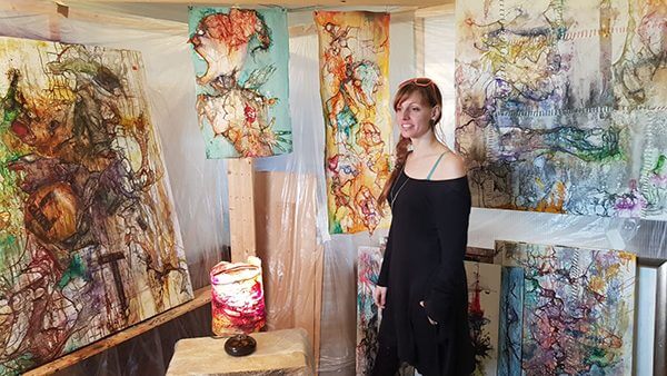 An artist stands with her mixed media abstract hanging paintings