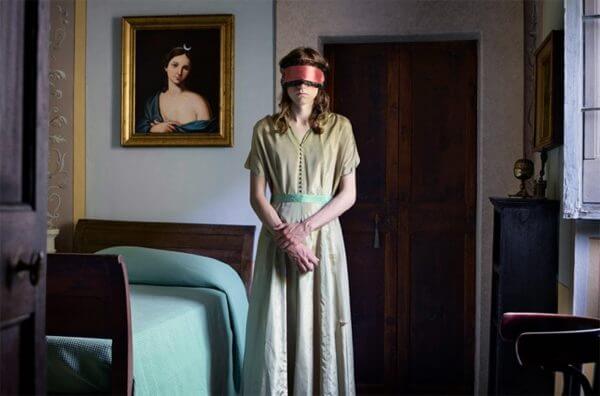 A photograph of a man in a blindfold in a room with a sleigh bed and painted portrait on the wall