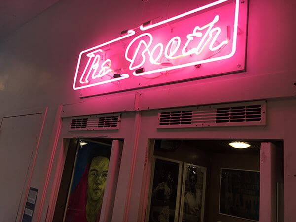 A pink neon sign spelling "The Booth" over glass doors to an art gallery