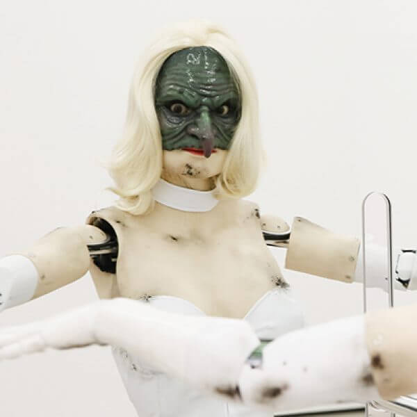 A sculpture of a humanoid robot wearing a blonde wig and grotesque half mask