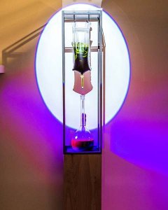 Tarah Rhoda, Ourglass, 2017. Spinach, ethanol, IV bag, volumetric flask, syringe, ultraviolet light, 10” x 10” x 73” Photos Credit-Scott McCullough