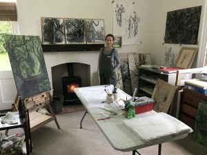 An artist in overalls standing in her studio among paintings of trees