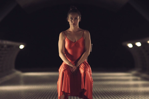 In a still from a short film, a dancer in a red dress sits on a chair.