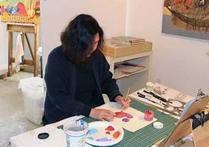 A painter sits at a table working on one of her paintings