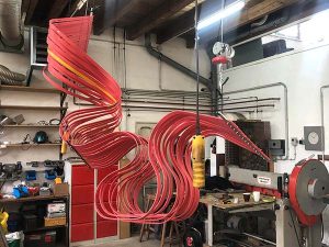 A metal sculpture in progress, with multiple curvy strands of red metal