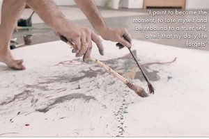 An image of an artist hovering two paint brushes over a canvas on the floor